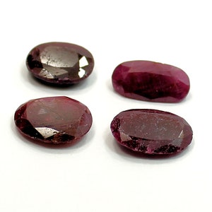 May include: Four oval-shaped, faceted gemstones with a deep red color. The gemstones are arranged in a row on a white surface.