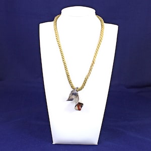 May include: A gold-colored rope necklace with a silver-toned pendant featuring a leaf design and a small, reddish-brown stone. The necklace is displayed on a white mannequin against a dark blue background.
