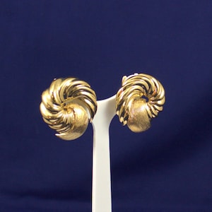 May include: A pair of gold-coloured clip-on earrings. Each earring has a spiral design with a textured, rounded base. The earrings are displayed on a white stand against a dark blue background.