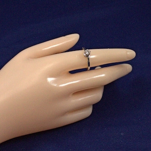 May include: A silver ring with a single, round, clear gemstone is shown on a finger. The ring is set against a dark blue background. The ring's band is thin and delicate.