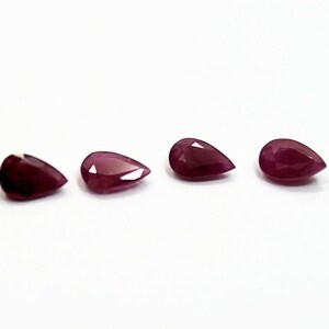 May include: Four pear-shaped, deep red gemstones are displayed against a white background. The gems have a polished, reflective surface, and their color is a rich, saturated hue. The arrangement is simple, with the stones evenly spaced.