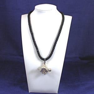 May include: A silver-toned flower pendant necklace with a black rope chain. The pendant is a detailed floral design with curved petals and small, round accents. The necklace is displayed on a white bust against a blue background.