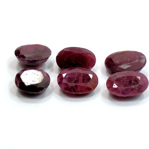 May include: Six oval-shaped, faceted ruby gemstones in various shades of red. The gemstones are arranged in a row on a white surface.