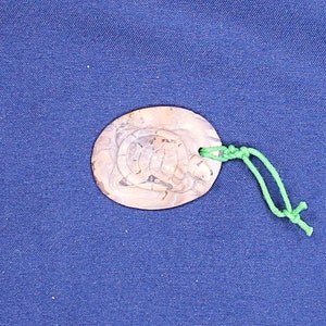 Peut inclure: Un pendentif ovale en bois brun avec un motif de tortue sculpté, suspendu à une ficelle verte.