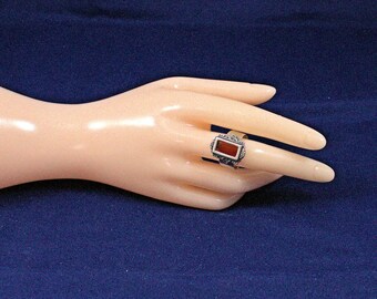 May include: A silver ring with a rectangular, reddish-brown stone. The ring has an ornate, detailed design around the stone. The ring is displayed on a mannequin hand against a dark blue background. The ring is a piece of jewelry.