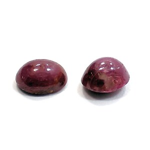 May include: Two oval-shaped cabochon gemstones, both a deep reddish-purple colour. The stones are smooth and polished, with a slight sheen.