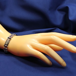 May include: A silver and gold bracelet with a Greek key design. The bracelet is on a mannequin's hand.