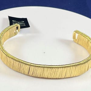 May include: A gold-toned bangle bracelet made of thin, stacked metal rings. The bracelet is open and has a simple design.