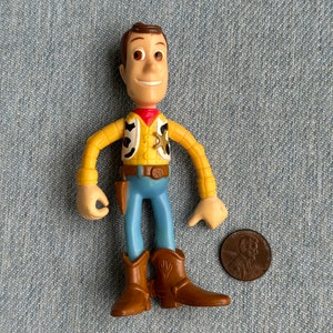 May include: A Woody action figure, featuring a yellow shirt, blue pants, and brown cowboy boots. The figure has a red neckerchief and a sheriff's badge. A penny is next to the figure for size comparison.