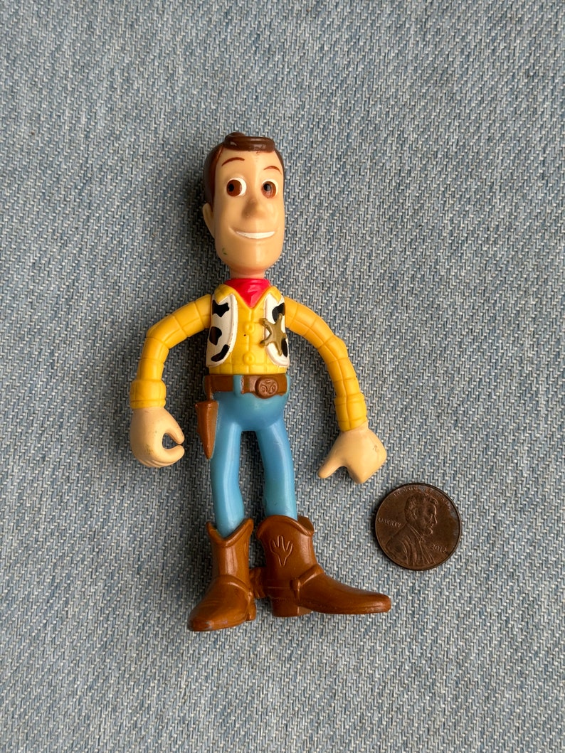 May include: A Woody action figure, featuring a yellow shirt, blue pants, and brown cowboy boots. The figure has a red neckerchief and a sheriff's badge. A penny is next to the figure for size comparison.