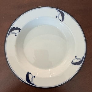 May include: White ceramic plate with a dark blue rim and decorative blue floral accents. The plate has a shallow bowl shape and is suitable for serving soups, salads, or other dishes. The design is simple and elegant.