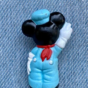 May include: A blue and white plastic figurine of Mickey Mouse dressed as a sailor. He is wearing a blue sailor hat, a red scarf, and blue overalls. He is holding a white paintbrush in his right hand.