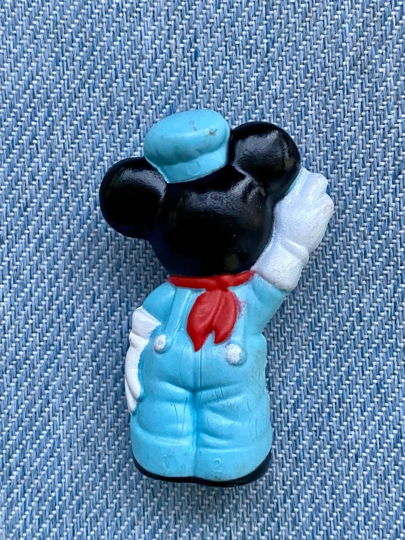 May include: A blue and white plastic figurine of Mickey Mouse dressed as a sailor. He is wearing a blue sailor hat, a red scarf, and blue overalls. He is holding a white paintbrush in his right hand.