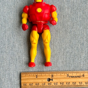 Może przedstawiać: Plastikowa figurka Iron Mana w kolorze czerwonym i ż&oacute;łtym, o wysokości około 10 cm. Figurka ma czerwoną płytę piersiową z ż&oacute;łtym kołem w środku oraz ż&oacute;łte nogi i ramiona. Figurka stoi na drewnianej linijce.