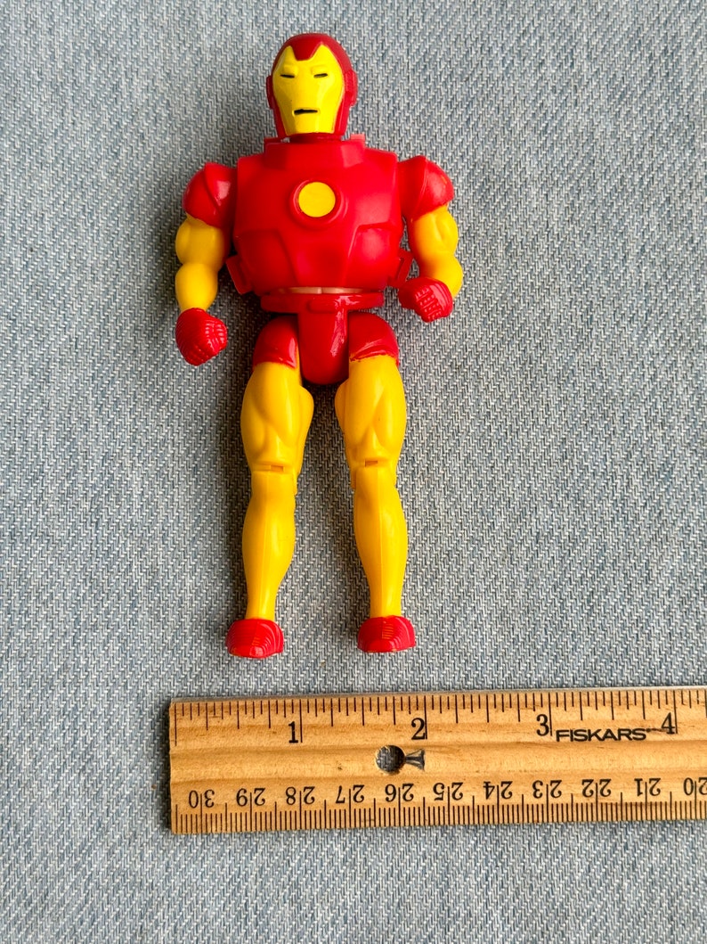 Może przedstawiać: Plastikowa figurka Iron Mana w kolorze czerwonym i ż&oacute;łtym, o wysokości około 10 cm. Figurka ma czerwoną płytę piersiową z ż&oacute;łtym kołem w środku oraz ż&oacute;łte nogi i ramiona. Figurka stoi na drewnianej linijce.