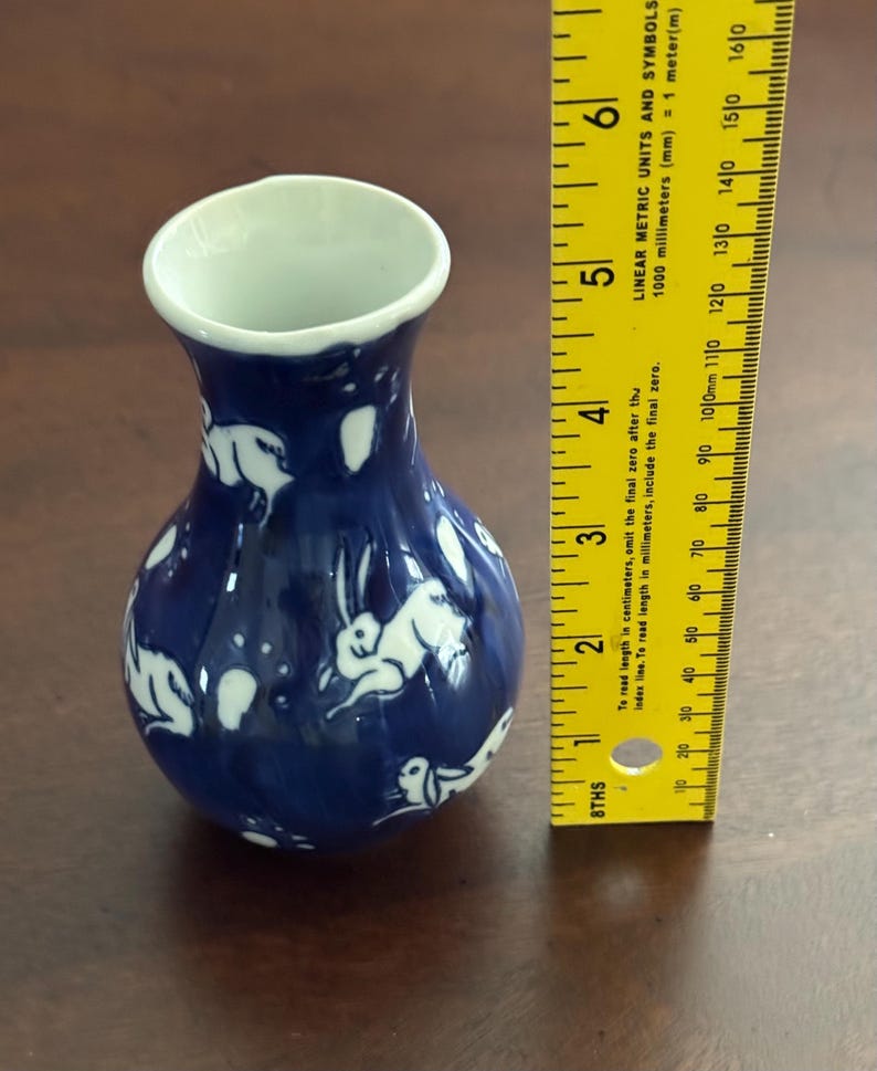 May include: A small blue and white ceramic vase with a design of white rabbits on a blue background. The vase is approximately 5 inches tall.