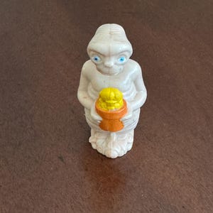 May include: A small, off-white figurine of the extraterrestrial character E.T. The figure has large blue eyes and is holding a yellow object in an orange container. The figurine is standing on a brown surface.