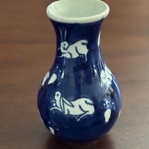 May include: A small, blue ceramic vase with a white glaze. The vase is decorated with a pattern of white rabbits.