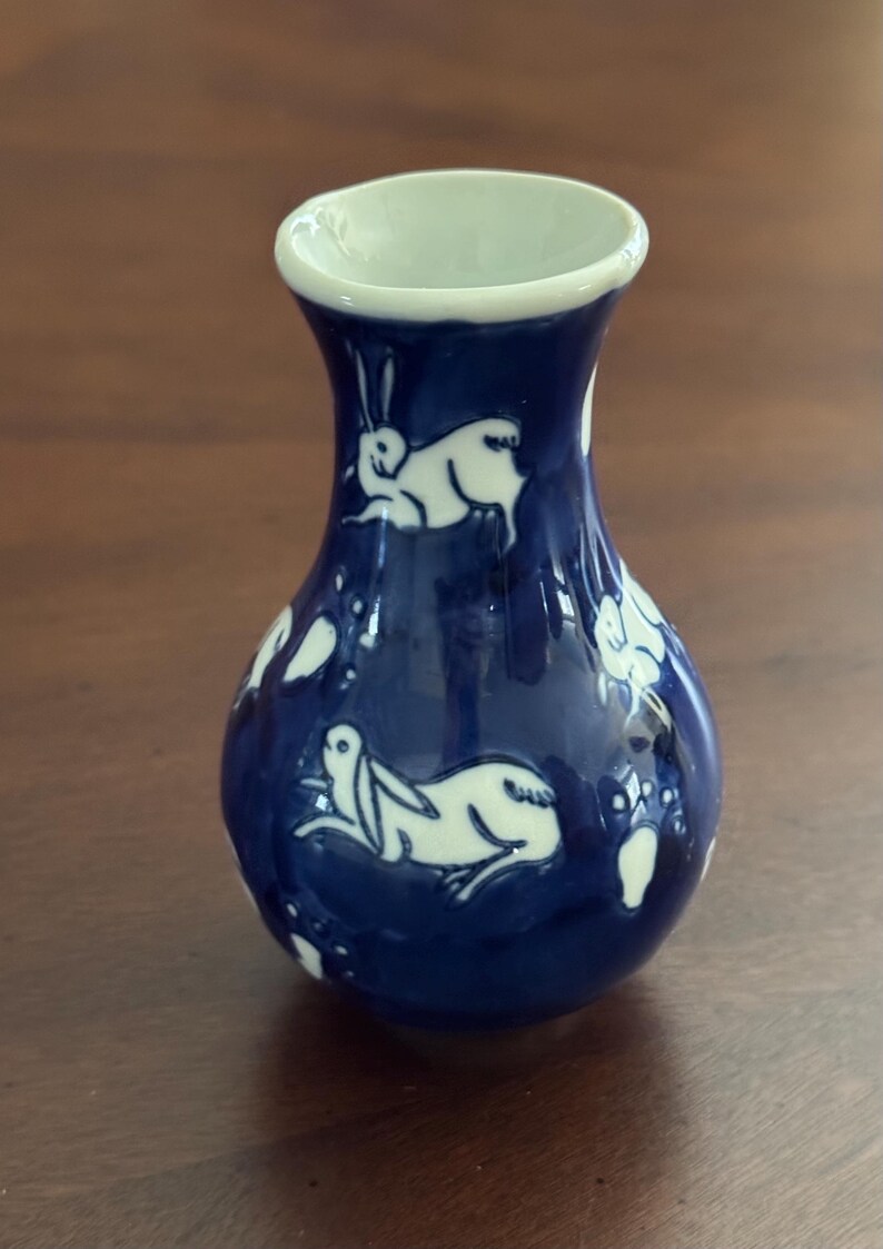 May include: A small, blue ceramic vase with a white glaze. The vase is decorated with a pattern of white rabbits.