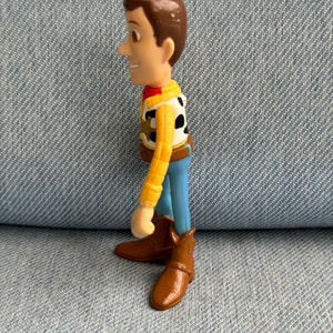 May include: A plastic figurine of a character from the animated film series Toy Story. The figure has brown hair, a yellow shirt, a black and white vest, blue pants, and brown cowboy boots. The figure is standing on a blue denim surface.