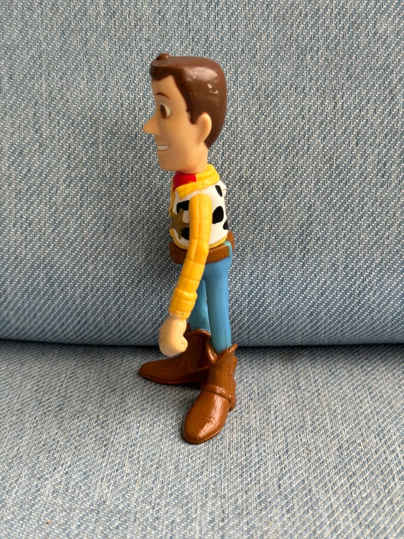 May include: A plastic figurine of a character from the animated film series Toy Story. The figure has brown hair, a yellow shirt, a black and white vest, blue pants, and brown cowboy boots. The figure is standing on a blue denim surface.