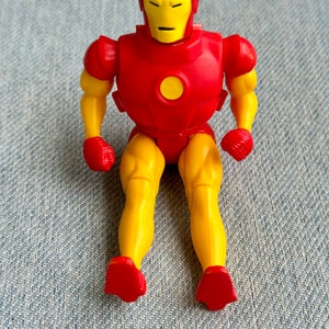 Może przedstawiać: Plastikowa figurka Iron Mana, superbohatera z komiks&oacute;w Marvela, w kolorze czerwonym i ż&oacute;łtym. Figurka siedzi ze skrzyżowanymi nogami i zgiętymi ramionami.