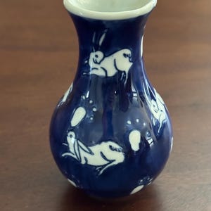 May include: A small blue ceramic vase with a white glaze. The vase is decorated with a pattern of white rabbits and their footprints.