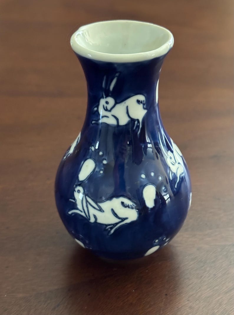 May include: A small blue ceramic vase with a white glaze. The vase is decorated with a pattern of white rabbits and their footprints.