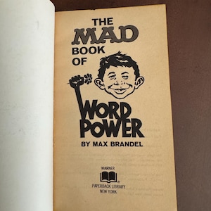 Pode incluir: Livro aberto com capa bege. O t&iacute;tulo &eacute; "THE MAD BOOK OF WORD POWER" com uma ilustra&ccedil;&atilde;o de um rosto sorridente e um punho levantado segurando uma flor. O autor &eacute; Max Brandel.