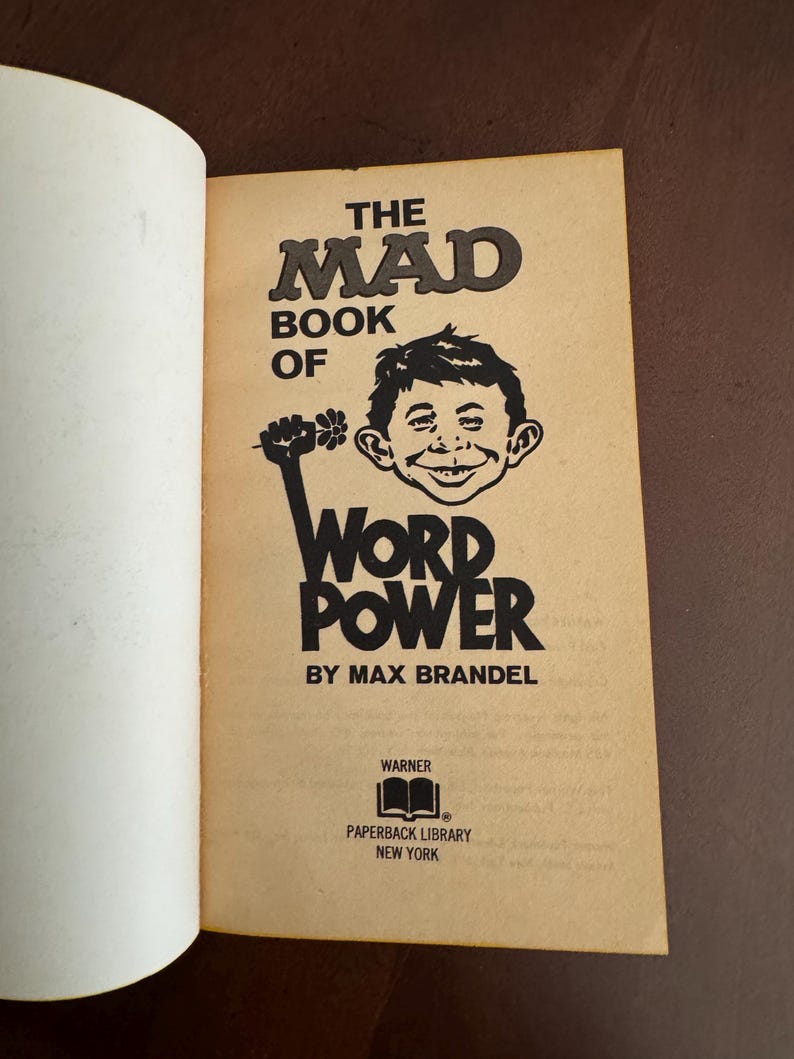 Pode incluir: Livro aberto com capa bege. O t&iacute;tulo &eacute; "THE MAD BOOK OF WORD POWER" com uma ilustra&ccedil;&atilde;o de um rosto sorridente e um punho levantado segurando uma flor. O autor &eacute; Max Brandel.