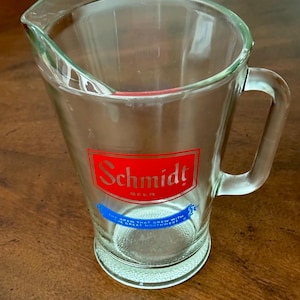 May include: A clear glass beer pitcher with a handle and a pouring spout. The pitcher has a red and white "Schmidt Beer" logo on the front. Below the logo is a blue banner with white text. The pitcher is sitting on a wooden surface.