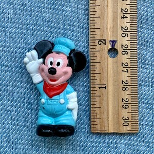 May include: A small plastic figurine of Mickey Mouse wearing a blue and white sailor outfit. He is waving with his right hand and has a red bandana around his neck. The figurine is about 1.5 inches tall.