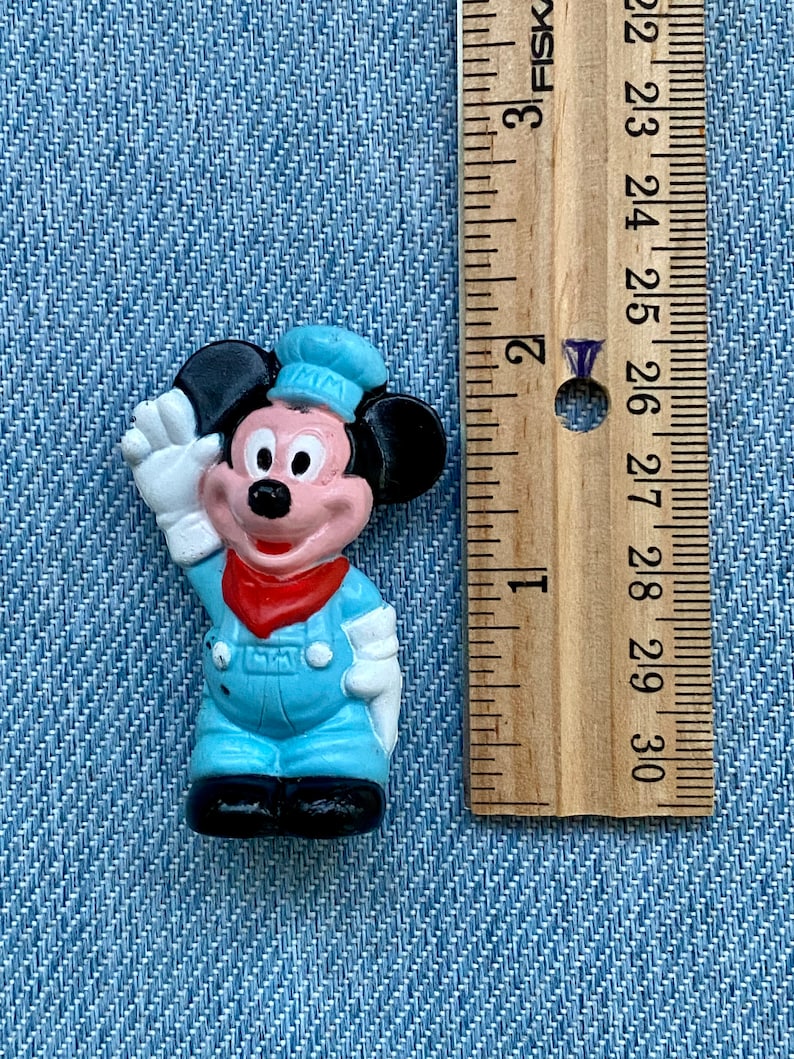 May include: A small plastic figurine of Mickey Mouse wearing a blue and white sailor outfit. He is waving with his right hand and has a red bandana around his neck. The figurine is about 1.5 inches tall.