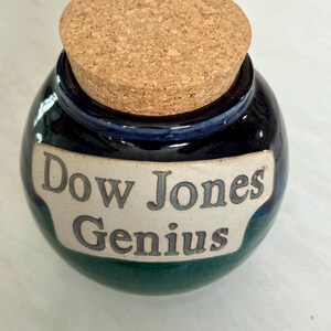 May include: A round ceramic jar with a cork lid. The jar is dark blue with a green base and a cream-colored label that reads "Dow Jones Genius" in gray lettering. The cork lid is light brown.