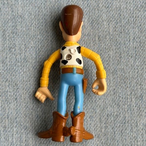 May include: A plastic toy figure of a cowboy character, viewed from the back. The figure has a brown head, yellow arms, a white vest with black spots, blue pants, and brown boots. A small holster is attached to the right side.