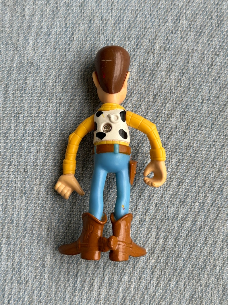 May include: A plastic toy figure of a cowboy character, viewed from the back. The figure has a brown head, yellow arms, a white vest with black spots, blue pants, and brown boots. A small holster is attached to the right side.