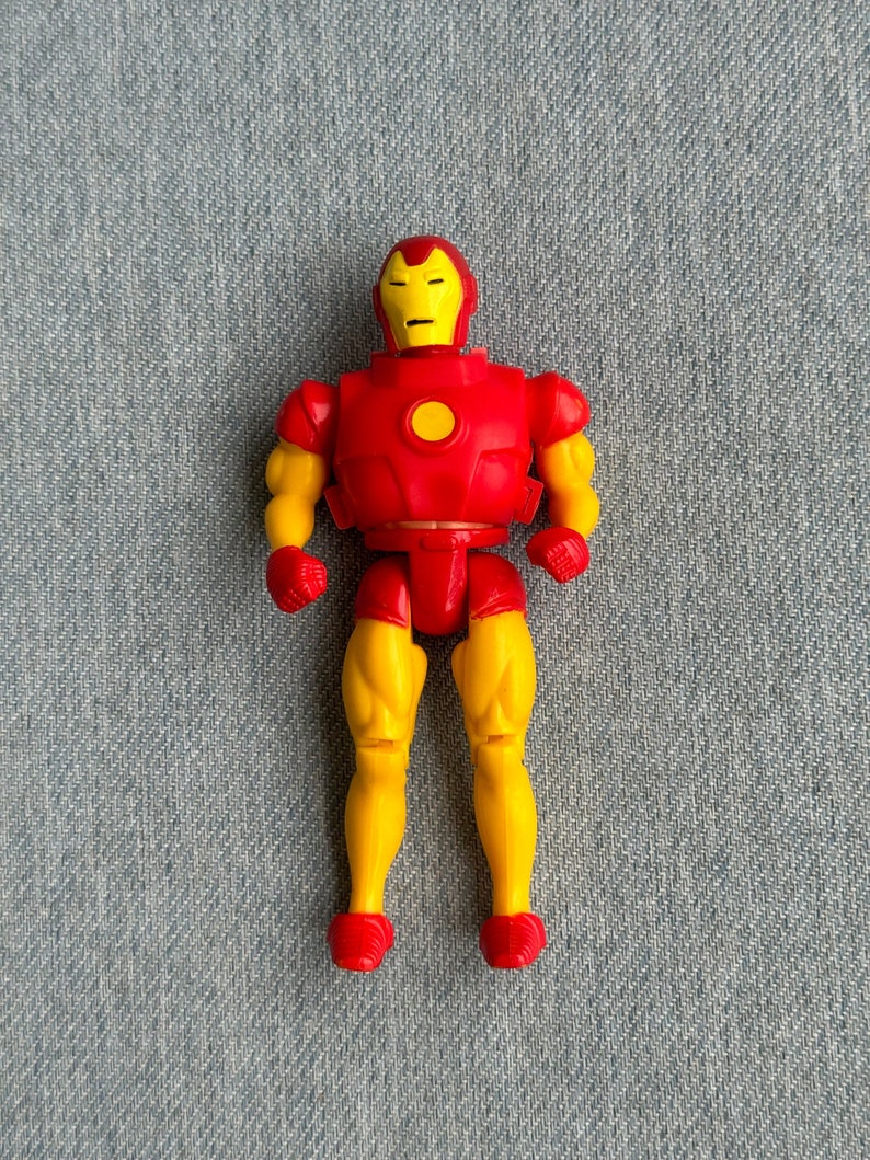 Może przedstawiać: Plastikowa figurka Iron Mana w kolorze czerwonym i ż&oacute;łtym. Figurka ma czerwoną płytę piersiową z ż&oacute;łtym kołem w środku. Figurka stoi na niebieskim dżinsowym tle.