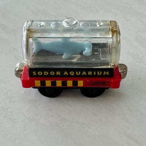 May include: A toy train car labeled "SODOR AQUARIUM" with a clear plastic aquarium containing a blue whale figurine. The train car is red with black wheels and silver accents.