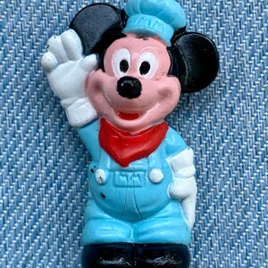 May include: A blue and black plastic figurine of Mickey Mouse wearing a blue cap and overalls. He is waving with his right hand and has a red bandana around his neck.