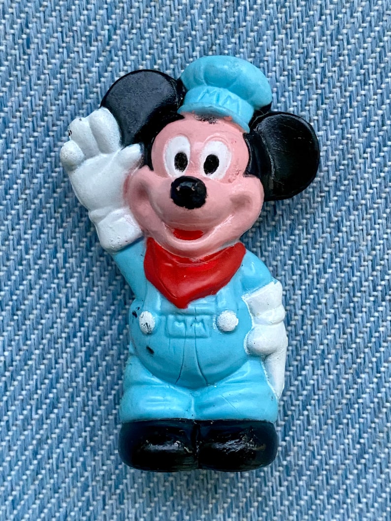 May include: A blue and black plastic figurine of Mickey Mouse wearing a blue cap and overalls. He is waving with his right hand and has a red bandana around his neck.