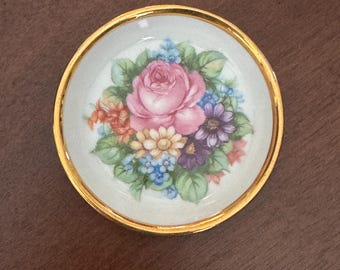 May include: Small, round decorative dish with a gold rim. The center features a floral design with a pink rose as the focal point, surrounded by blue, purple, and orange flowers and green leaves. The dish is set against a dark brown background.