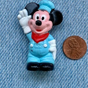 May include: A small plastic figurine of Mickey Mouse wearing a blue cap and overalls. He is waving with his right hand and has a red bandana around his neck.