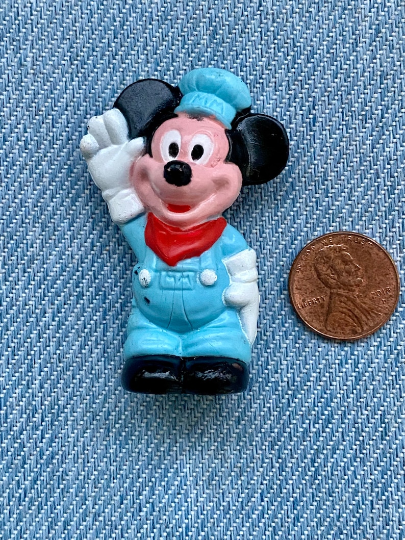May include: A small plastic figurine of Mickey Mouse wearing a blue cap and overalls. He is waving with his right hand and has a red bandana around his neck.