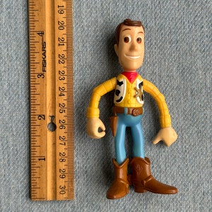 May include: A Woody action figure, featuring a yellow shirt, blue pants, and brown boots. The figure has a red neckerchief and a sheriff's badge. A wooden ruler is placed next to the figure, measuring up to 12 inches.