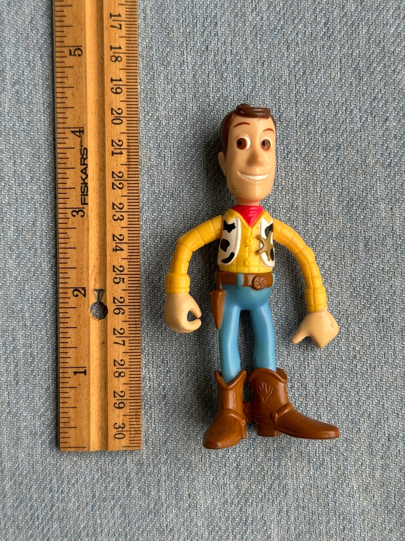 May include: A Woody action figure, featuring a yellow shirt, blue pants, and brown boots. The figure has a red neckerchief and a sheriff's badge. A wooden ruler is placed next to the figure, measuring up to 12 inches.