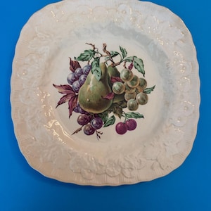 May include: A square ceramic plate with a cream-colored base and a scalloped edge featuring an embossed leaf pattern. The center of the plate displays a colorful illustration of a green pear surrounded by clusters of purple and green grapes.
