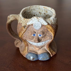 May include: A ceramic mug shaped like a woman's face with a smiling expression. The mug has brown and white glaze with blue eyes and a white 'RN' on the forehead.