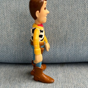 May include: A side profile of a Woody action figure from the Toy Story franchise. The figure has brown hair, a yellow shirt, a black and white vest, blue pants, and brown boots. The figure is standing on a denim surface.