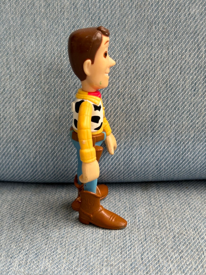 May include: A side profile of a Woody action figure from the Toy Story franchise. The figure has brown hair, a yellow shirt, a black and white vest, blue pants, and brown boots. The figure is standing on a denim surface.