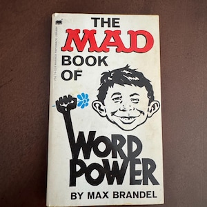 Pode incluir: Um livro de bolso vintage intitulado "The Mad Book of Word Power" de Max Brandel. A capa apresenta uma ilustra&ccedil;&atilde;o de desenho animado de um rosto sorridente e um punho erguido segurando uma flor. O t&iacute;tulo &eacute; em vermelho e preto.